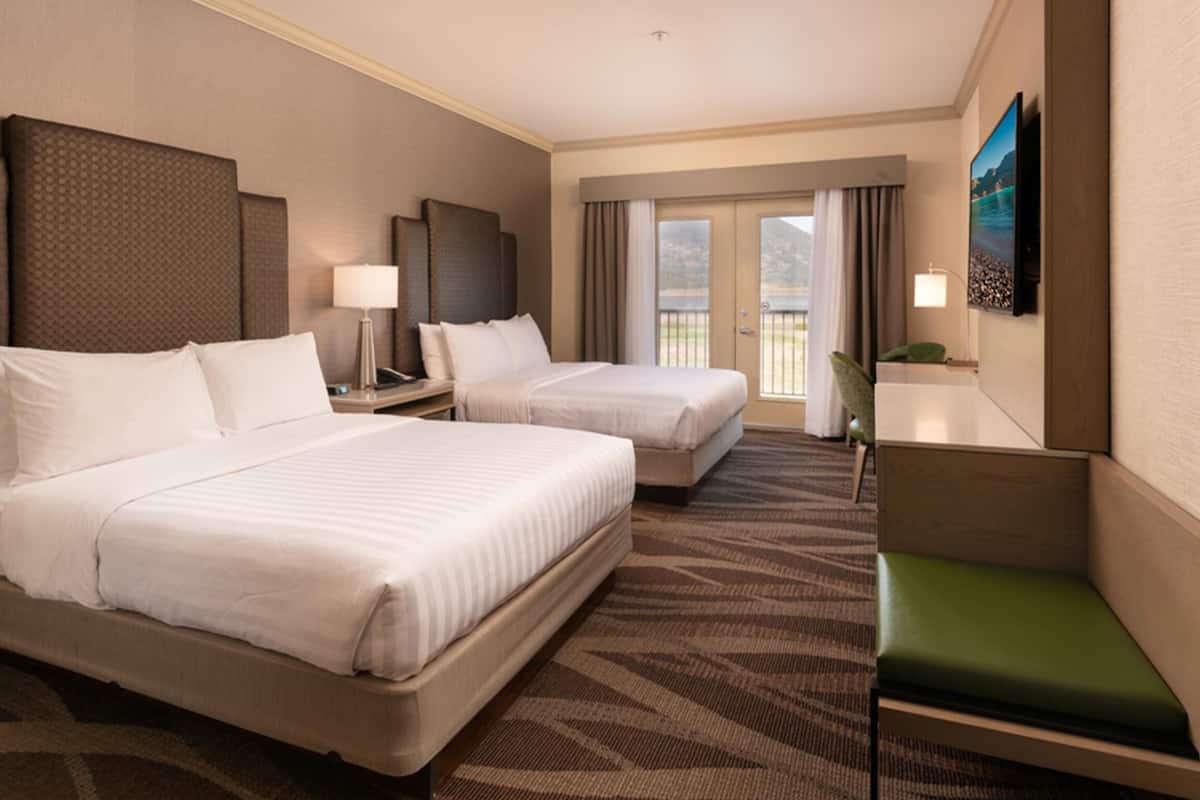 A hotel room with two beds, a green bench, a TV, and a balcony with curtains.