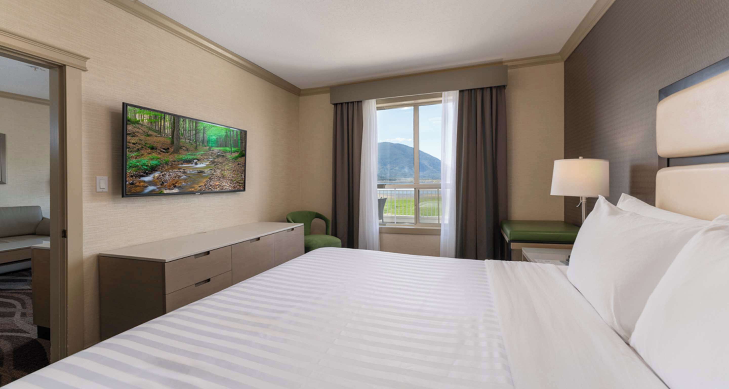 Una habitación de hotel con una cama grande, un televisor de pantalla plana montado en la pared, un ventanal con vistas a montañas y una silla verde.
