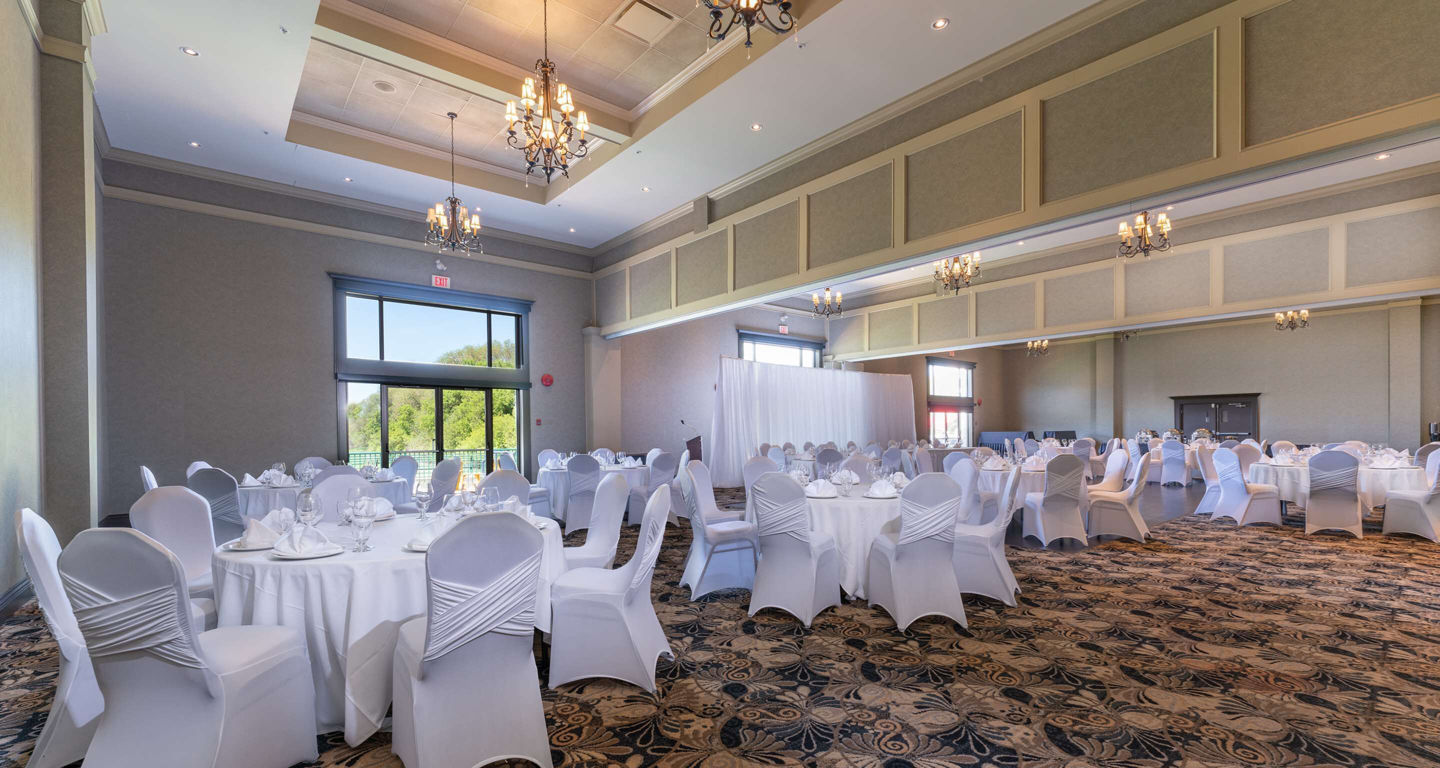 A banquet hall with round tables, white chairs, chandeliers, and a patterned carpet.