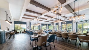 Un restaurant spacieux avec de hauts plafonds, des poutres apparentes et de grandes fenêtres offrant une vue sur l’extérieur. Les tables sont accompagnées de chaises et le sol est en bois.