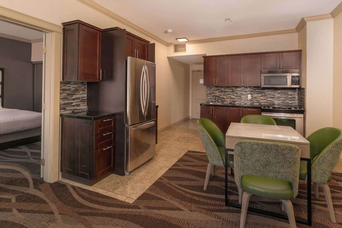 A compact hotel room with a kitchenette, a dining area with green chairs, and a bed with a white headboard.