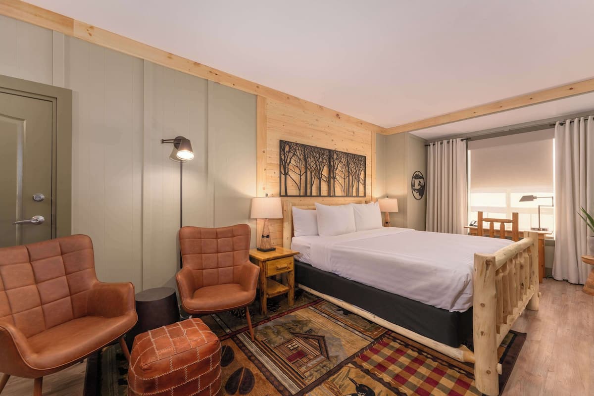 A hotel room with a bed, two armchairs, a nightstand, a lamp, a wall art piece, and a window with curtains.