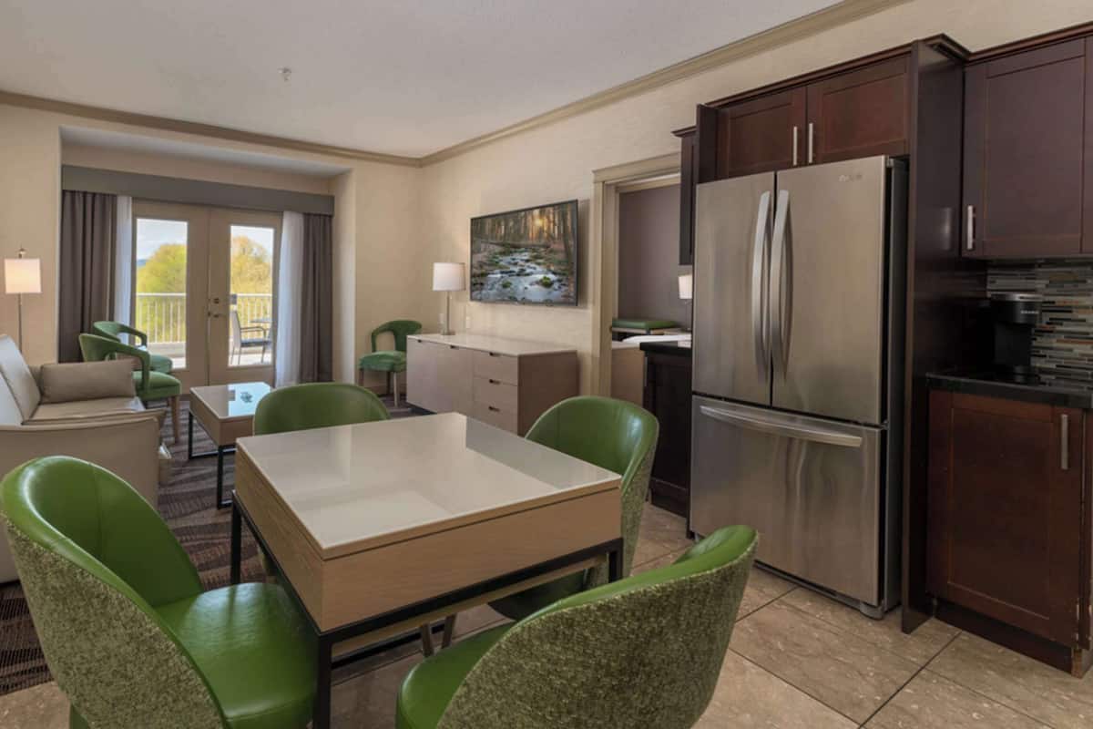 A hotel room with a dining area, a sofa, a table, green chairs, a kitchen with a refrigerator, and a painting on the wall.