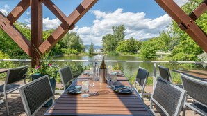 Un espace de restauration en plein air avec une table en bois dressée pour deux personnes, offrant une vue sur une rivière paisible et une végétation luxuriante.