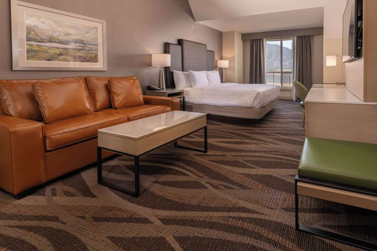 A hotel room with a brown leather sofa, a white coffee table, a green chair, a bed with white linens, a desk with a lamp, and a framed picture on the wall.