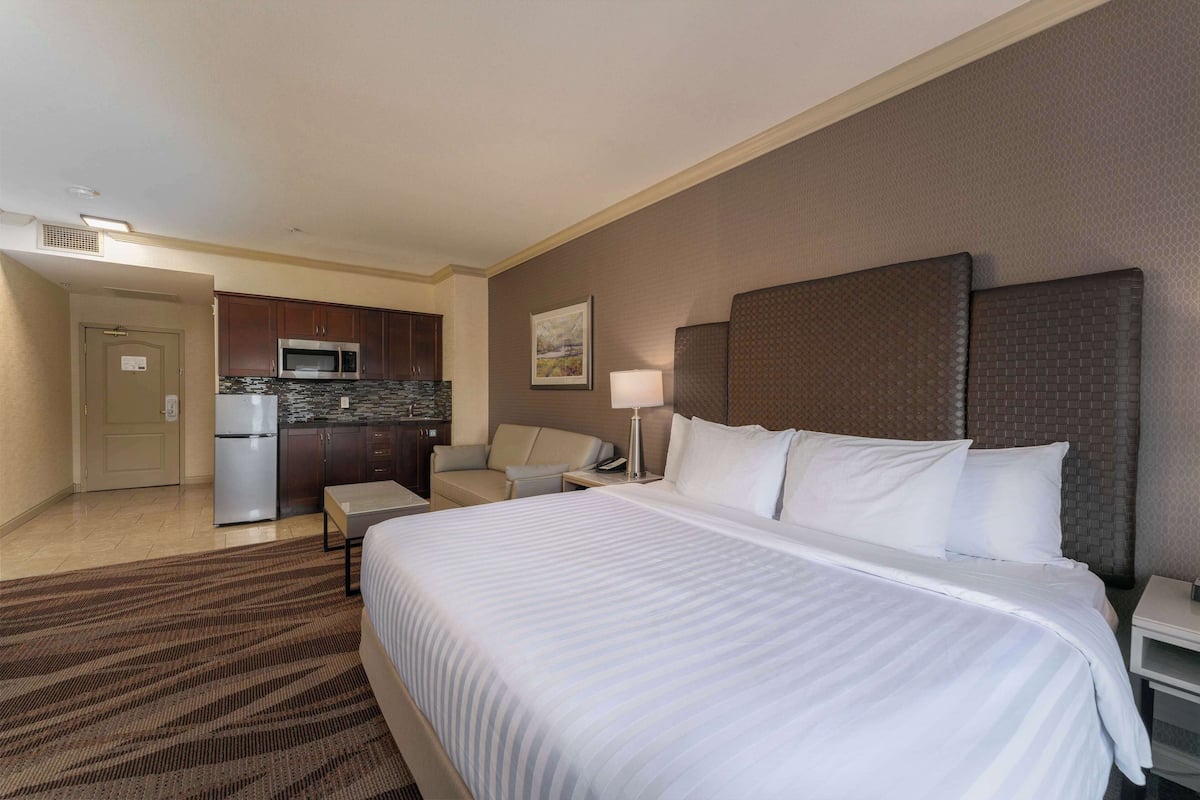 A hotel room with a large bed, a desk, a chair, a microwave, and a refrigerator.