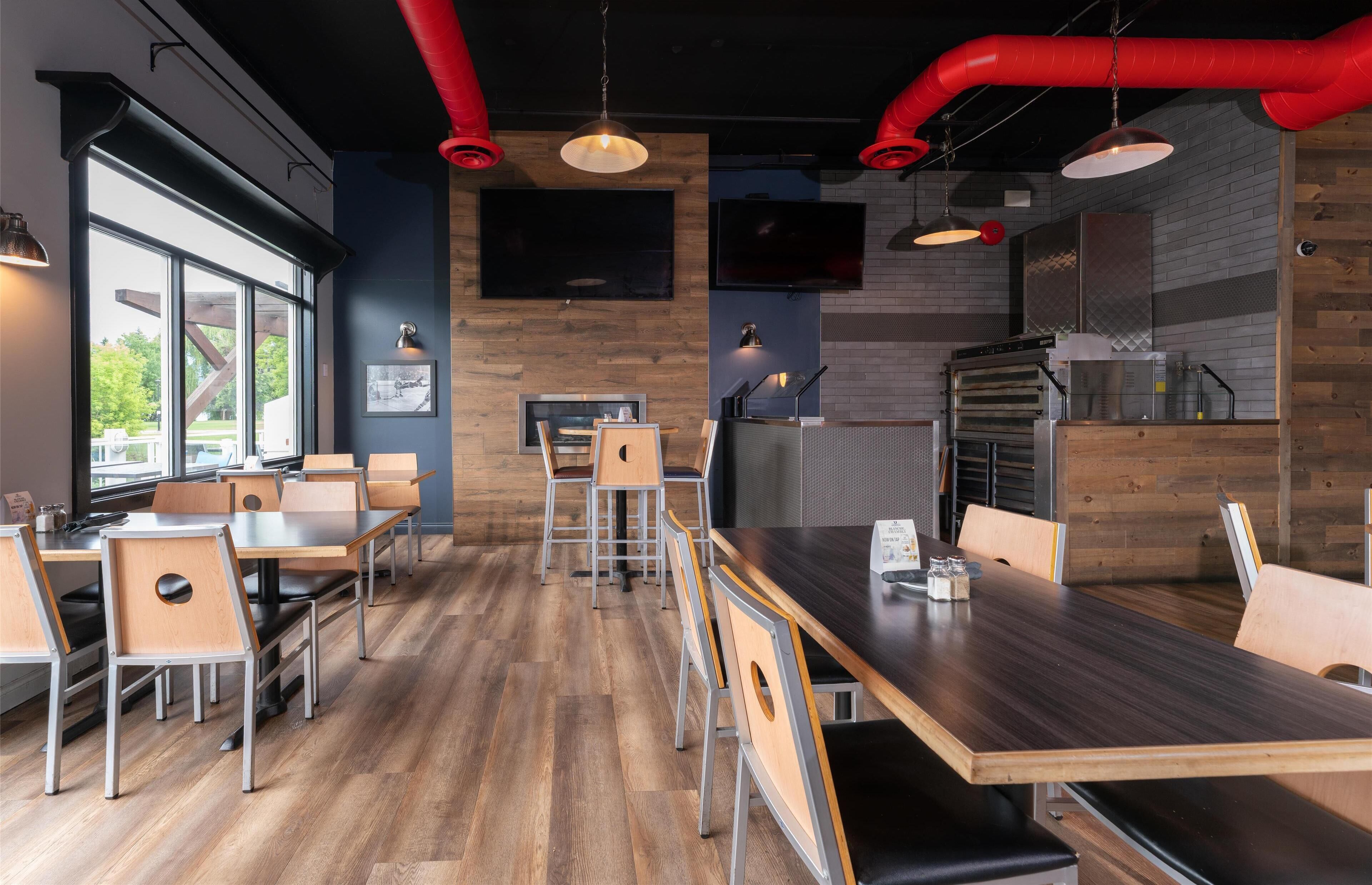 A modern restaurant interior with wooden flooring, metal chairs, and tables. There are large windows on one side, a kitchen area with a pizza oven, and a flat-screen TV mounted on the wall.