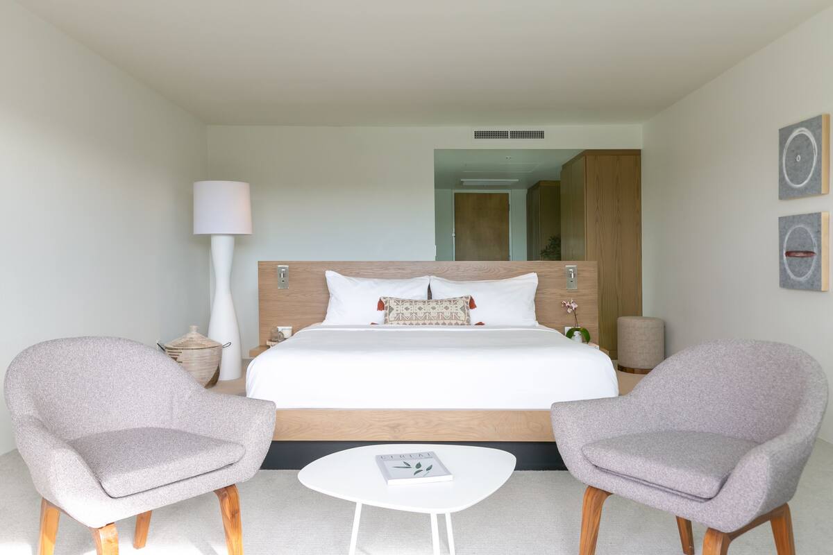 A modern hotel room with a large bed, two armchairs, a small white table, and a lamp.