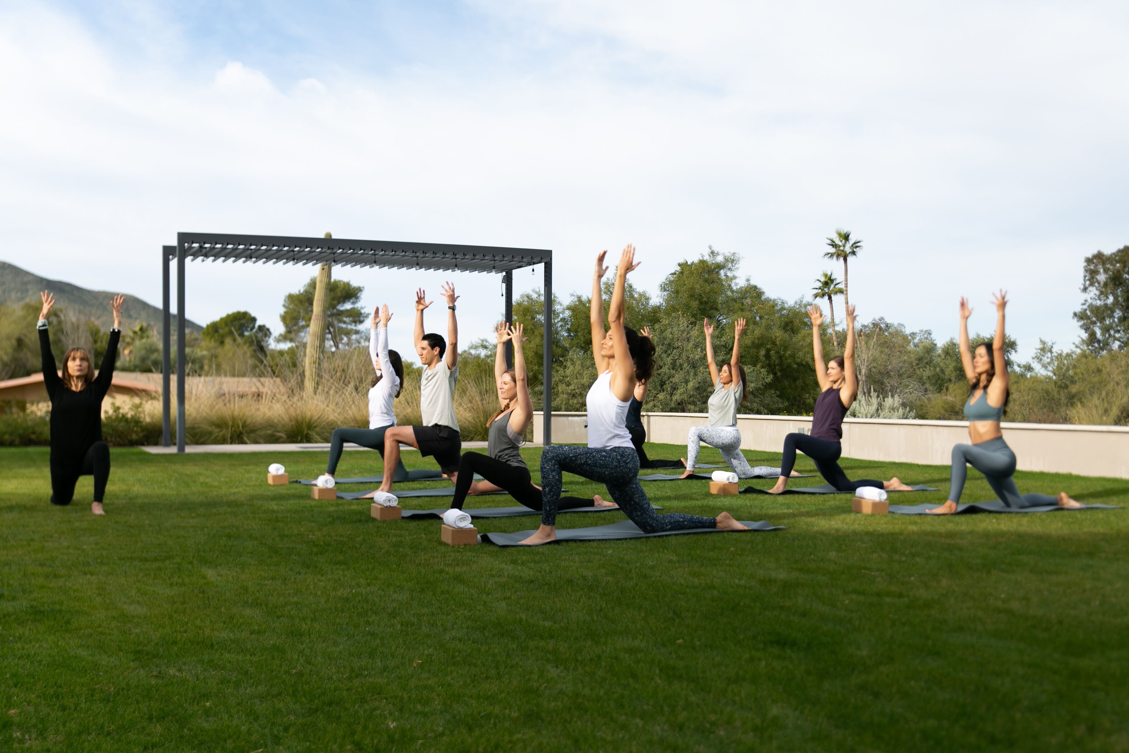 Un groupe de personnes pratiquant le yoga sur des tapis à l’extérieur, avec une pergola en arrière-plan.
