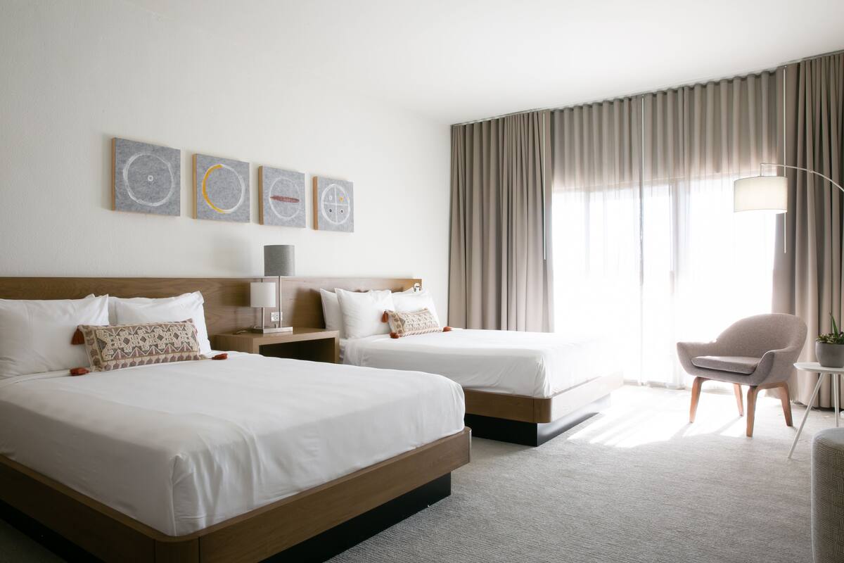 A hotel room with two beds, a chair, a lamp, and artwork on the wall.