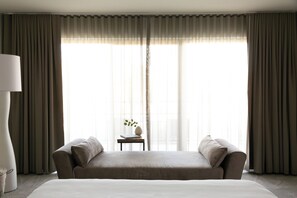 A hotel room with a large bed, a sofa, a small table with a vase, and sheer curtains.