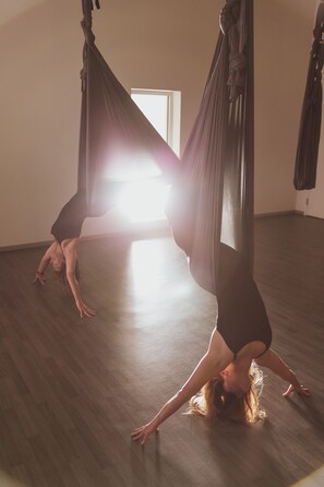 A person performing aerial yoga in a room with wooden flooring and a large window.