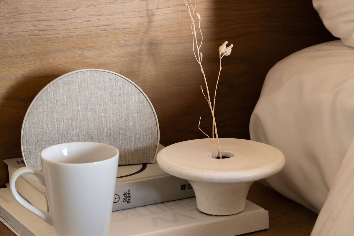 A neatly arranged bedside table with a white mug, a small white vase with dried branches, and a book titled "NEVVARCHITEK."