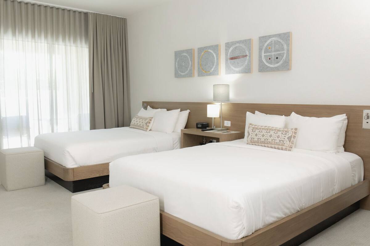 A hotel room with two beds, a bedside table with a lamp, and a large window with curtains.