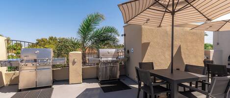 An outdoor patio with a grill, dining table, chairs, and umbrellas.