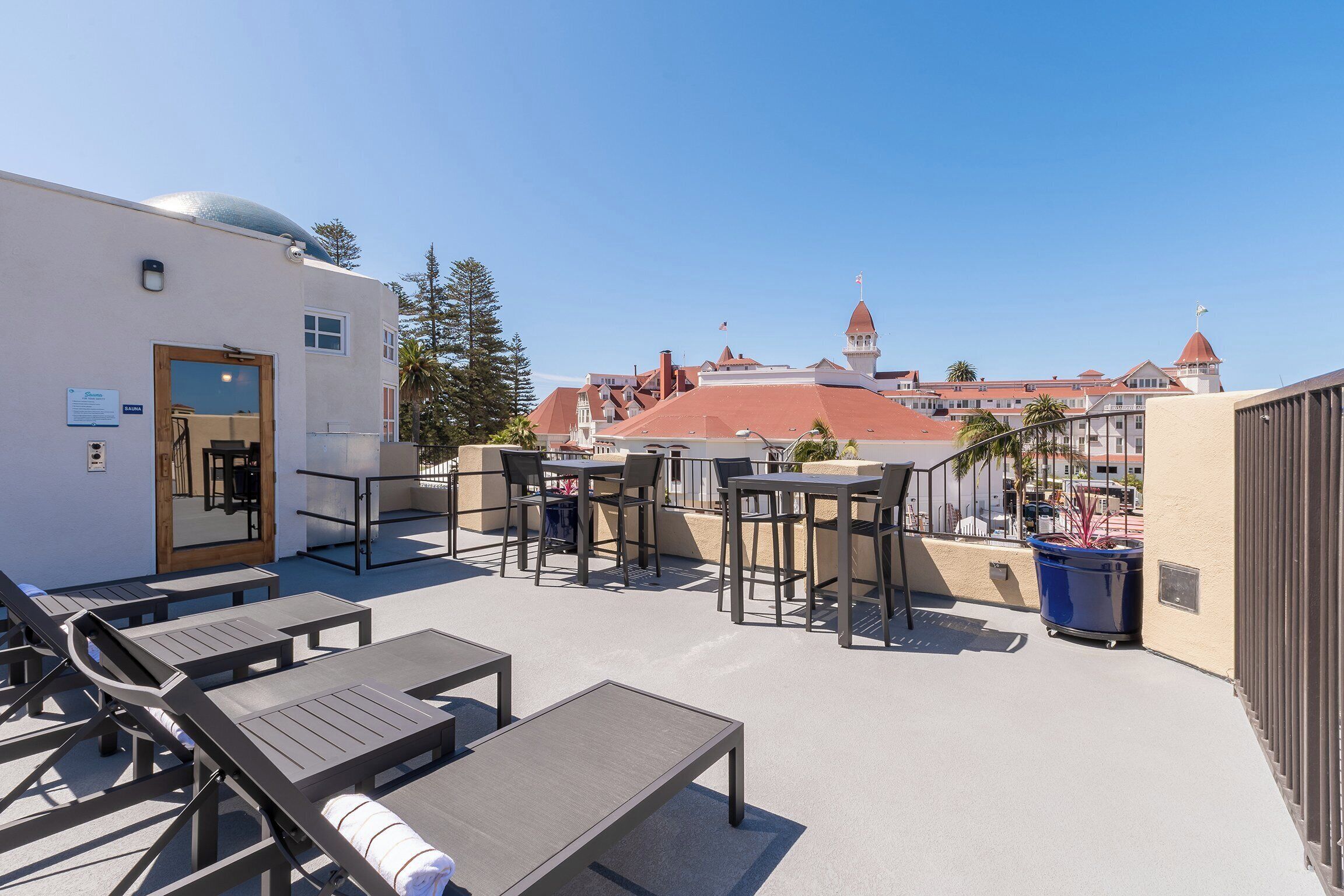 A rooftop terrace with lounge chairs and tables, a door, and a view of a historic building with a red roof.