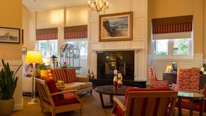 A well-lit living room with a fireplace, comfortable seating, and large windows with red valances. - Coronado Beach Resort (Coronado)
