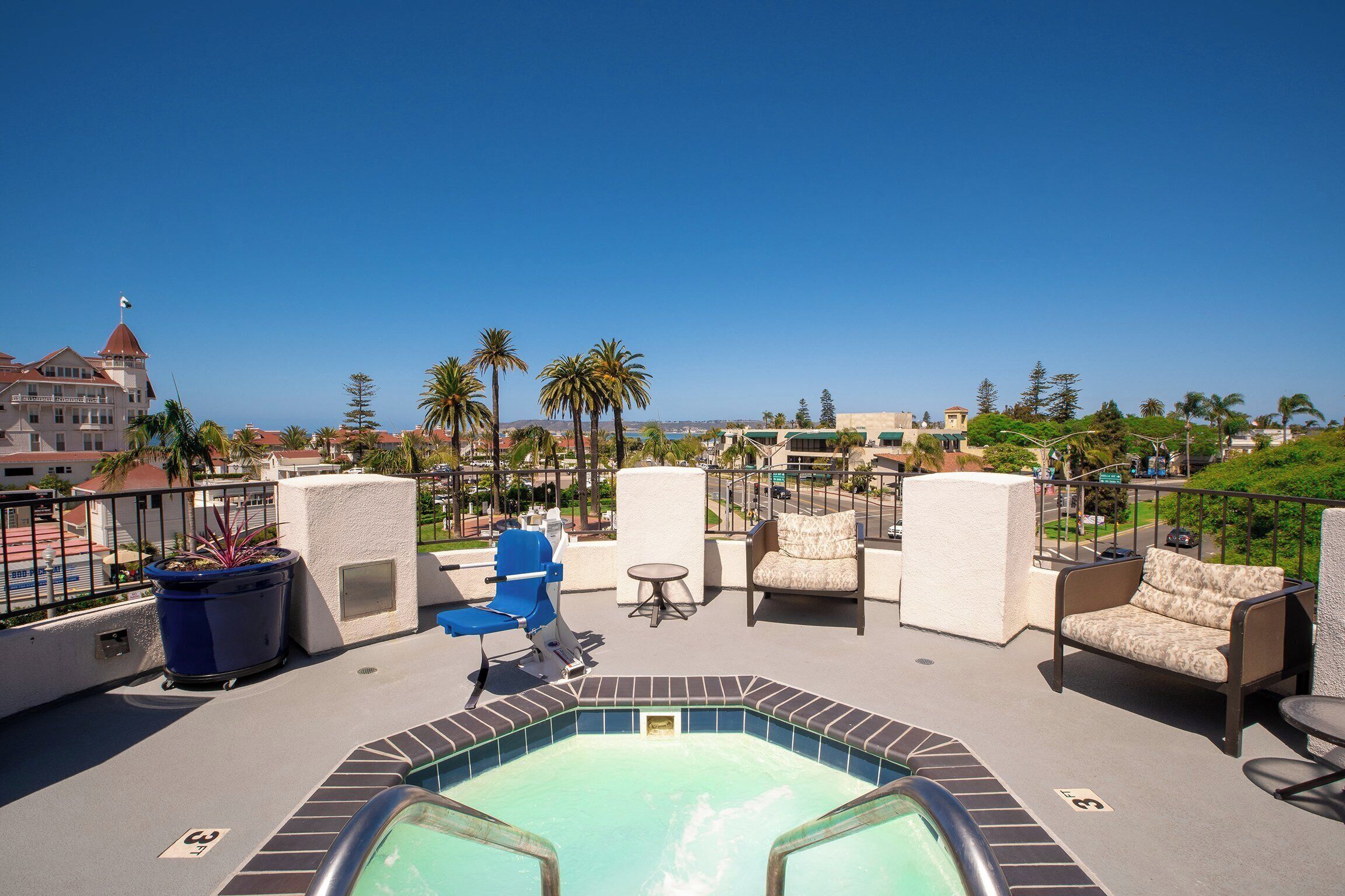 A rooftop pool area with lounge chairs and a hot tub, overlooking a cityscape with palm trees and buildings.