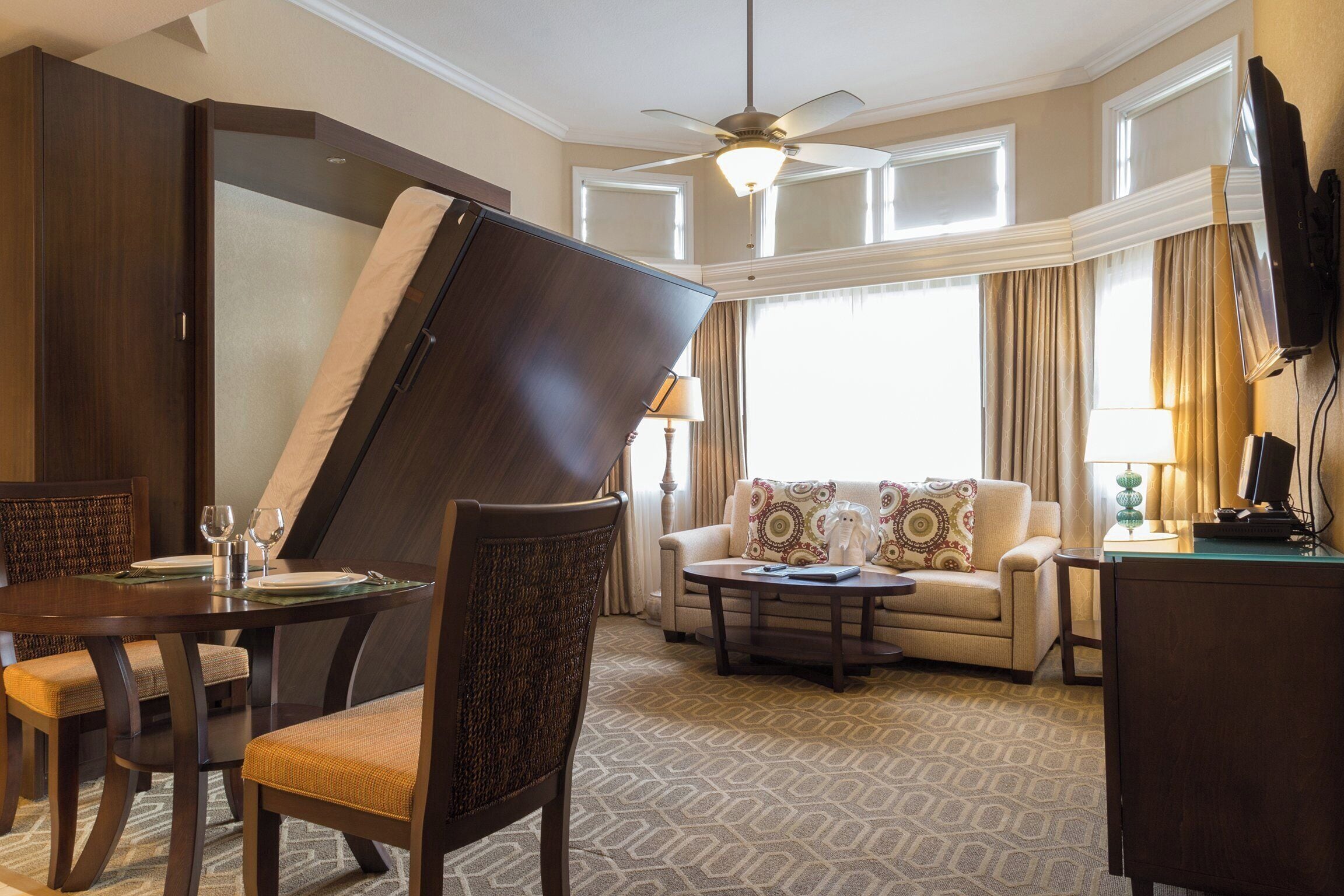 A hotel room with a dining area, a sofa, a TV, and a ceiling fan.
