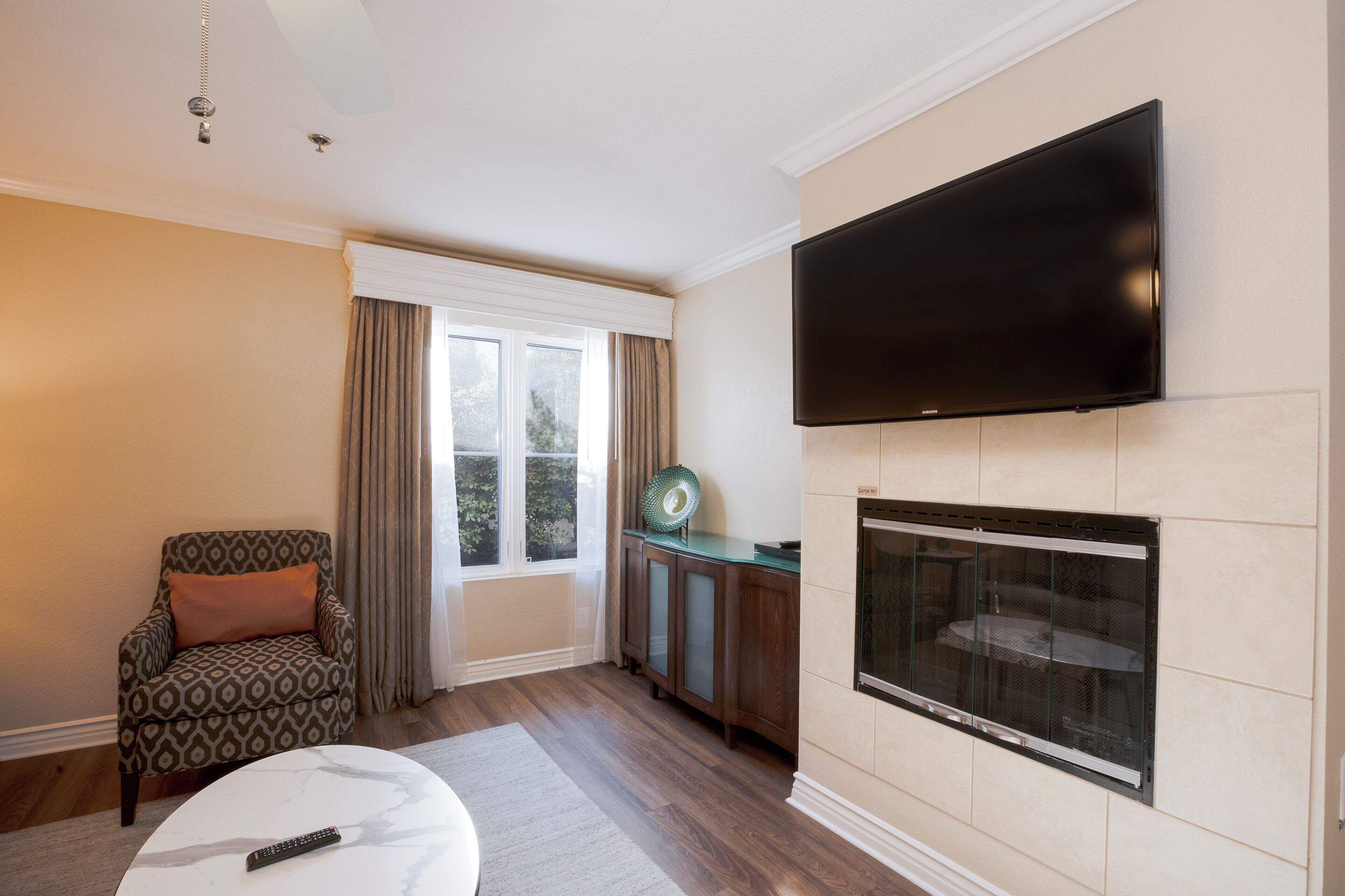 A living room with a flat-screen TV mounted on the wall, a fireplace, a patterned armchair, and a round coffee table.