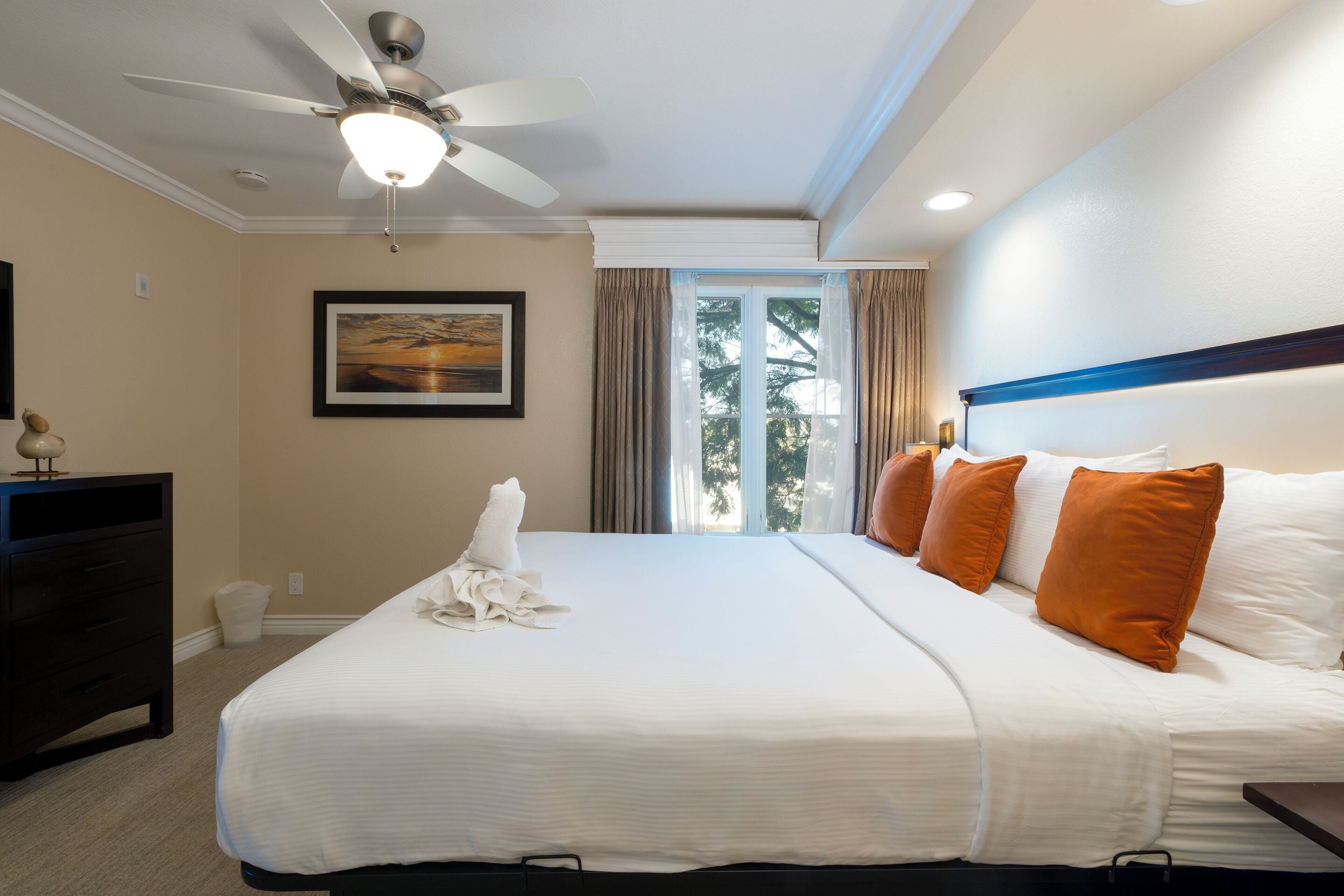 Habitación de hotel con una cama grande, un ventilador de techo, una ventana con cortinas, un cuadro enmarcado en la pared y un aparador.