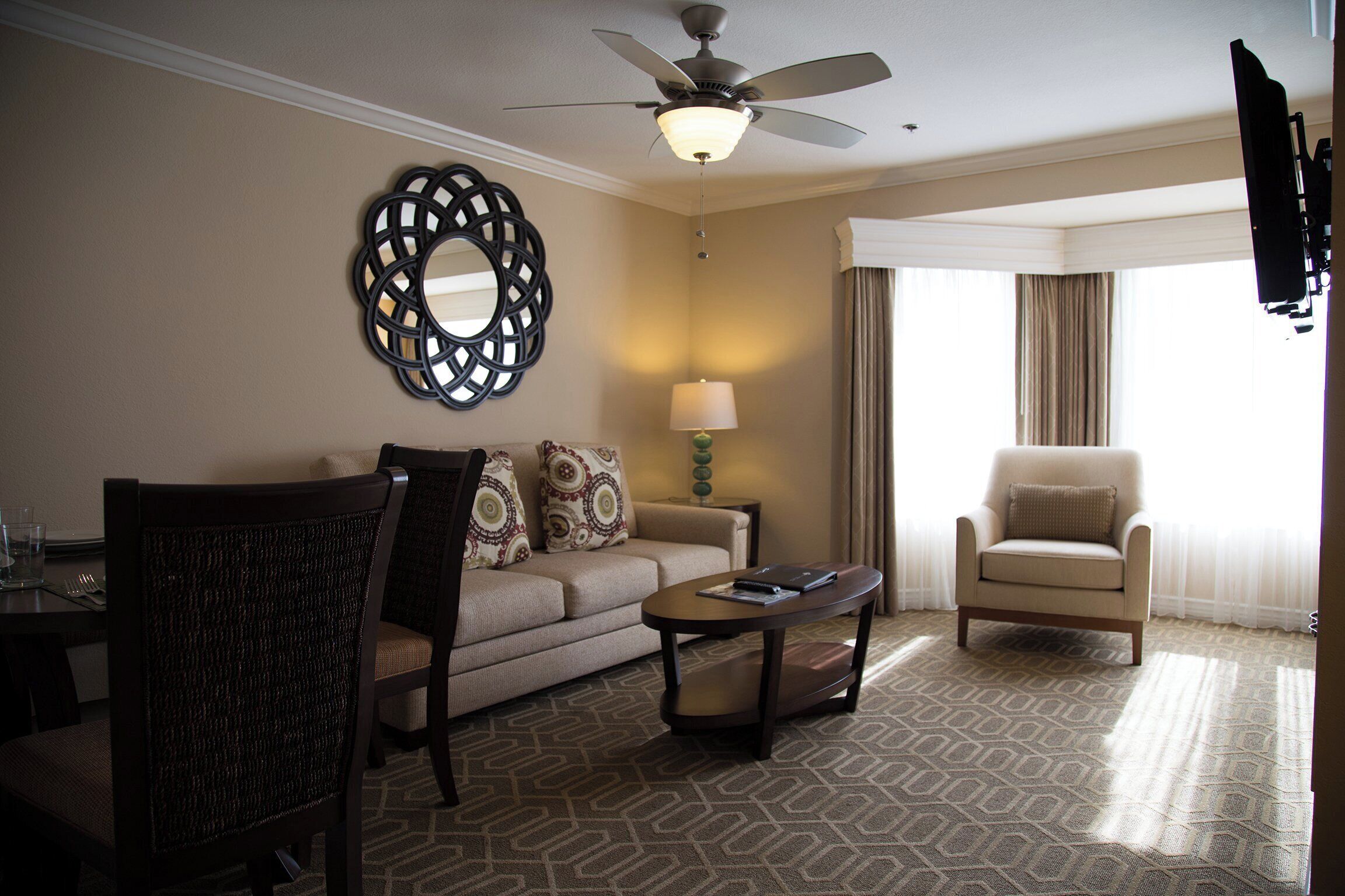 Habitación de hotel con sofá, sillón y una mesita de centro.  Tiene un ventilador de techo, una lámpara y un espejo decorativo en la pared.