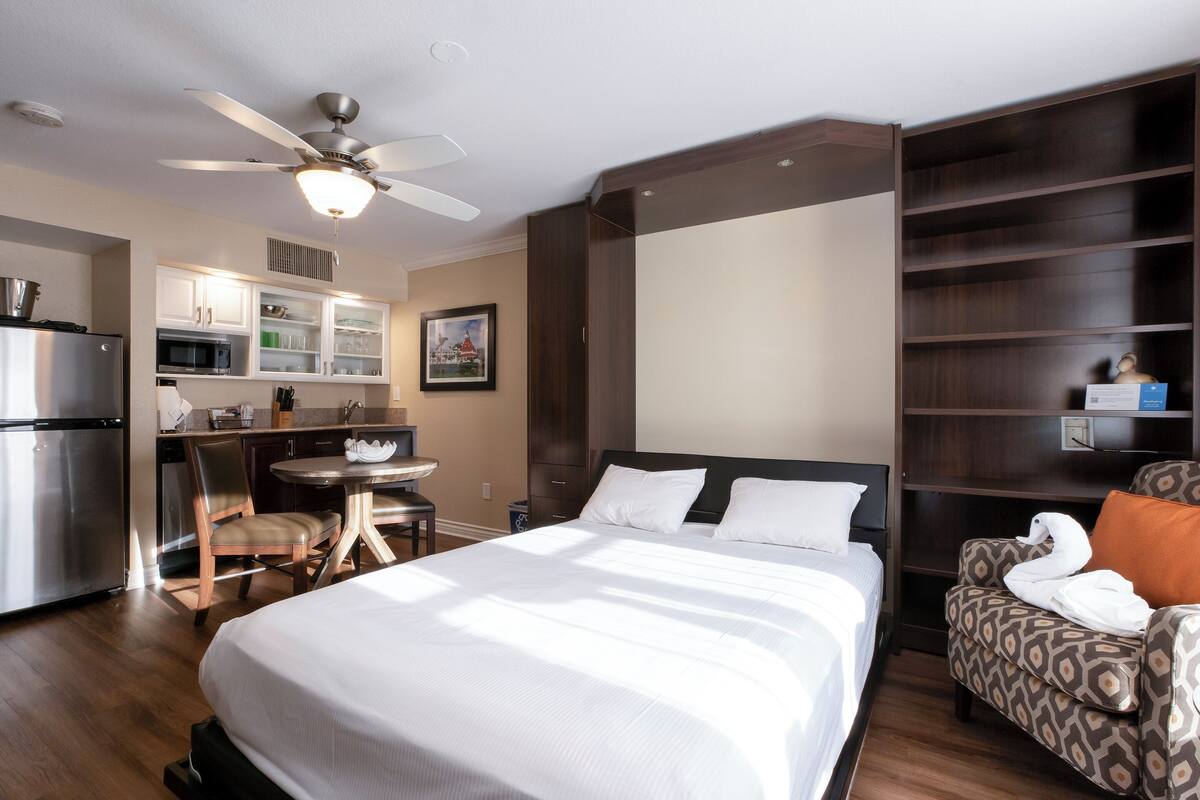 A hotel room with a bed, a dining area with a table and chairs, a kitchenette, and a ceiling fan.