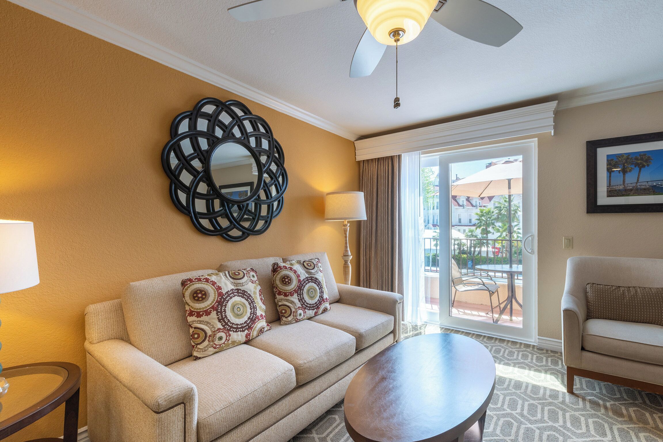 A hotel room with a beige sofa, a round coffee table, a ceiling fan, a large mirror, and a view of the outdoors through a sliding glass door.