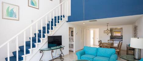 Una sala de estar con un sofá azul, una mesa de centro de vidrio y un televisor sobre un mueble. Se observa una escalera con escalones azules y barandillas blancas.