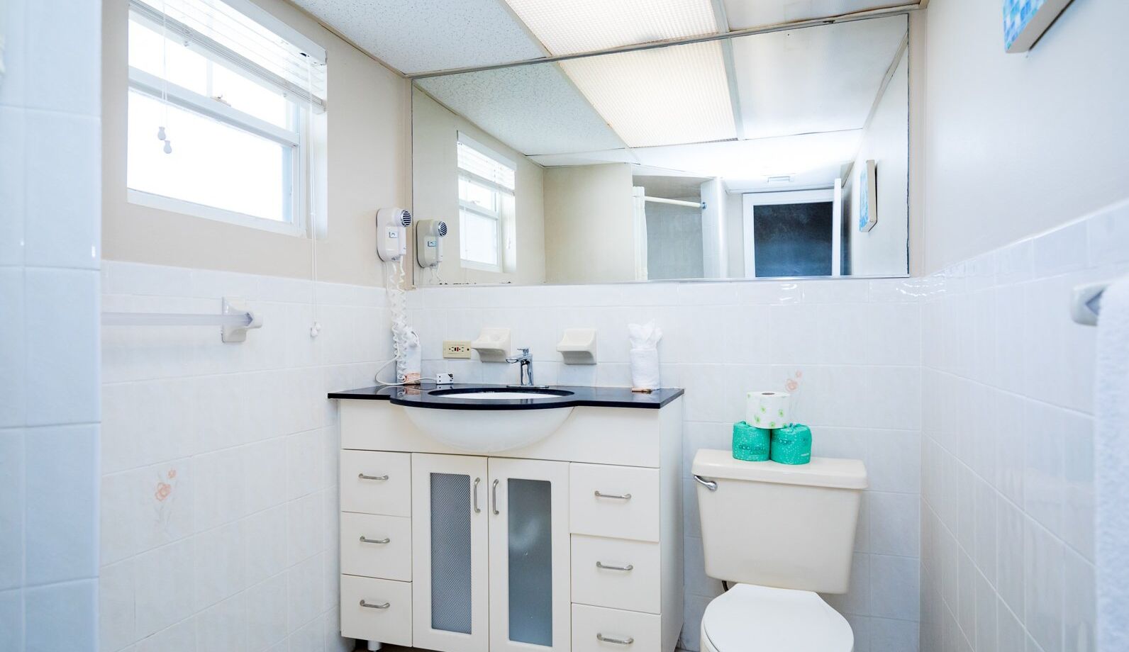 A bathroom with a white sink, mirror, and toilet. There are two rolls of toilet paper and two bottles of hand soap on the toilet tank.