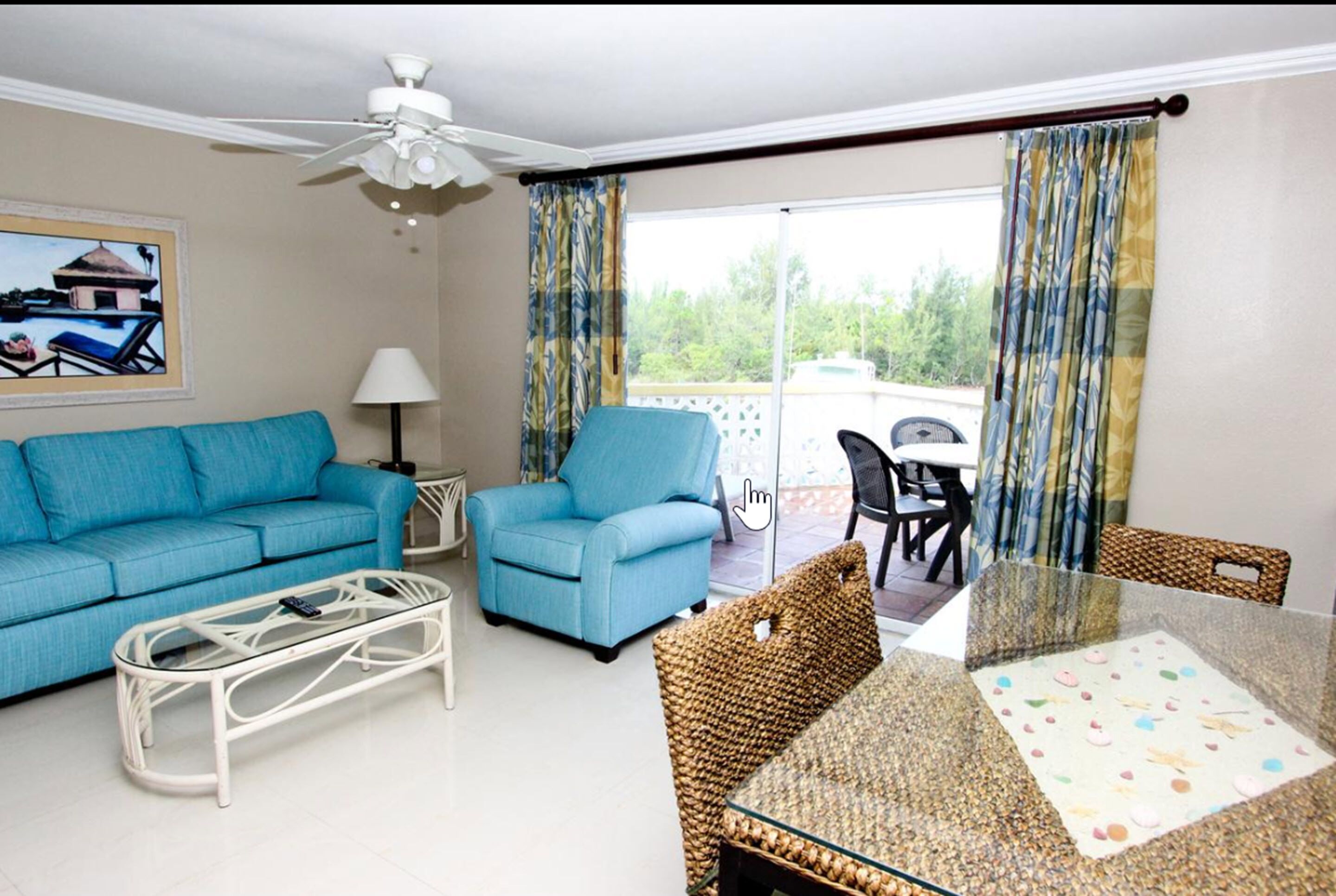 Uma sala de estar com sofá, poltrona, mesa de centro e uma mesa de jantar com toalha de mesa estampada. Há um ventilador de teto, um abajur e um quadro na parede.