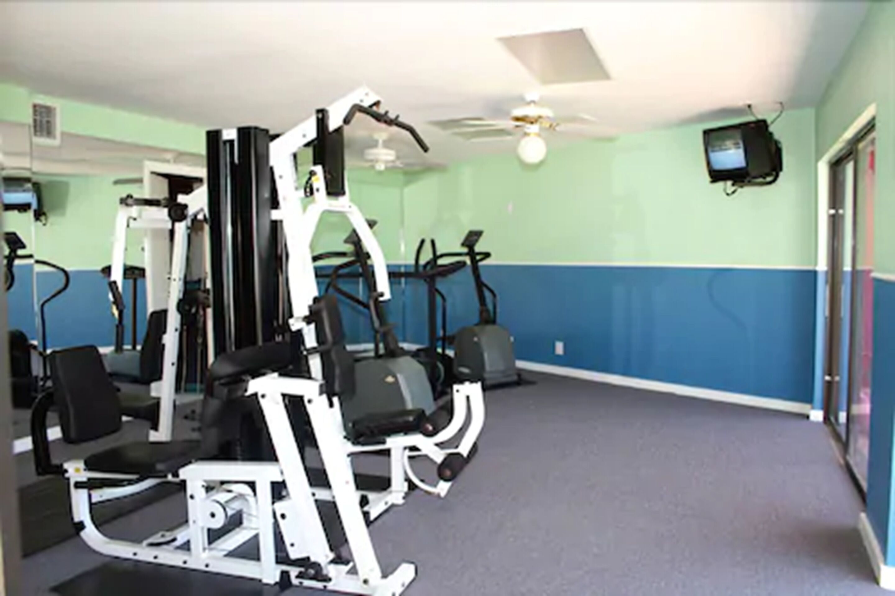 A gym with various exercise machines, a ceiling fan, and a television mounted on the wall.