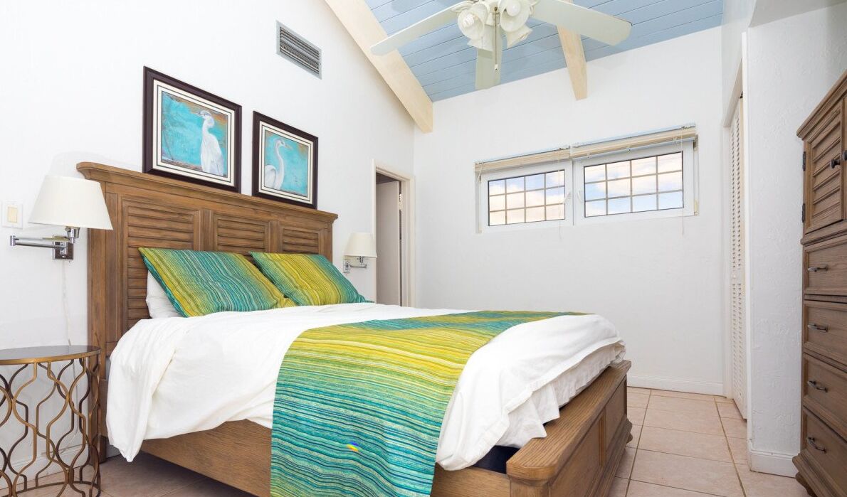 Quarto com cama de madeira, mesa de cabeceira, ventoinha no teto e duas imagens emolduradas na parede.