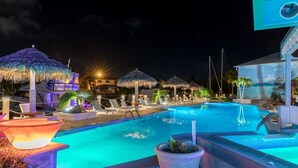 Ein Poolbereich bei Nacht mit Liegestühlen, Reetdach-Sonnenschirmen und einer großen beleuchteten Schautafel.