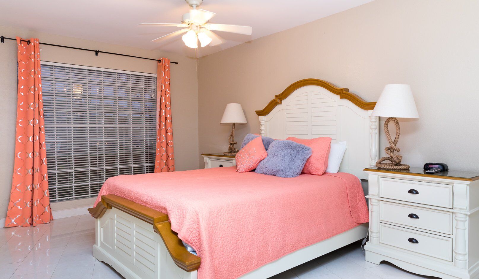 Une chambre à coucher comprenant un lit, deux tables de chevet, un ventilateur de plafond et une fenêtre avec des stores.