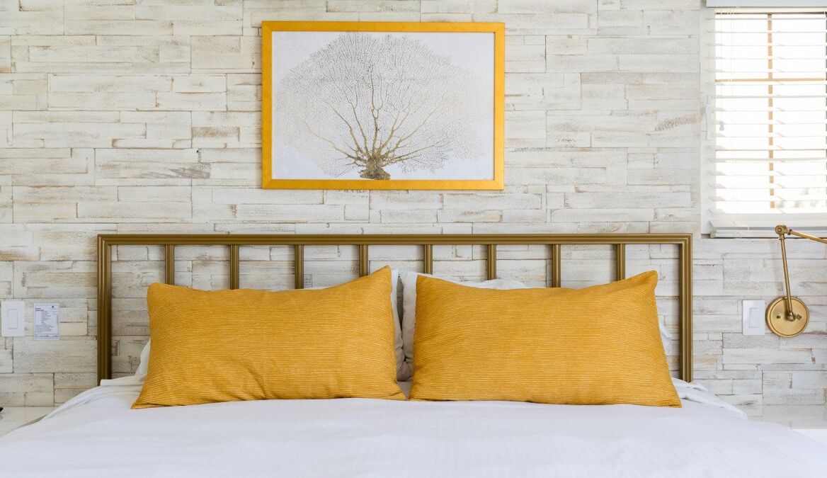 Un lit avec deux oreillers de couleur moutarde, une tête de lit en bois et une œuvre d’art représentant un arbre, encadrée, accrochée à un mur de briques.