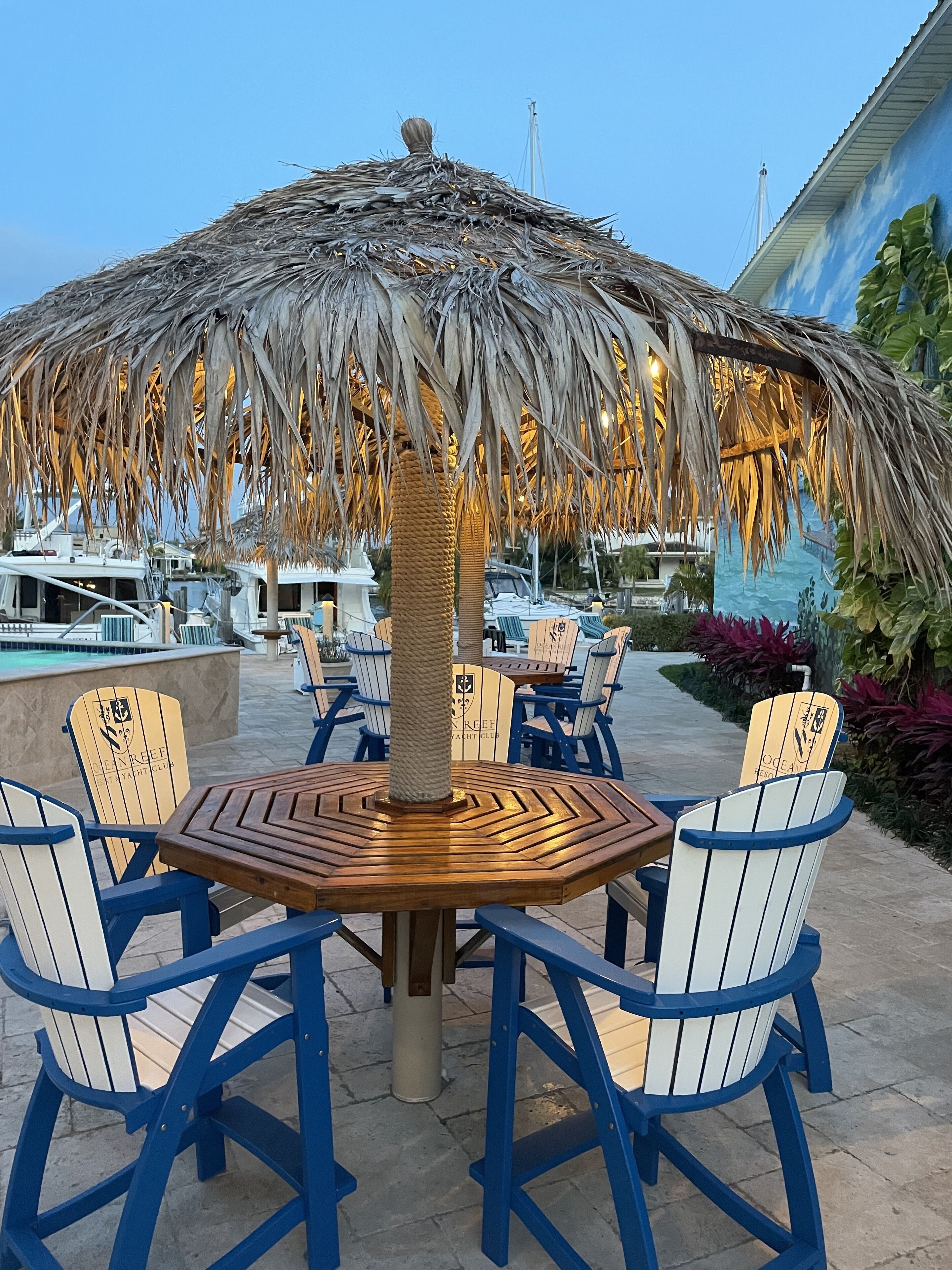 A round wooden table with blue and white chairs, a thatched umbrella, and a poolside setting with a view of a bridge.