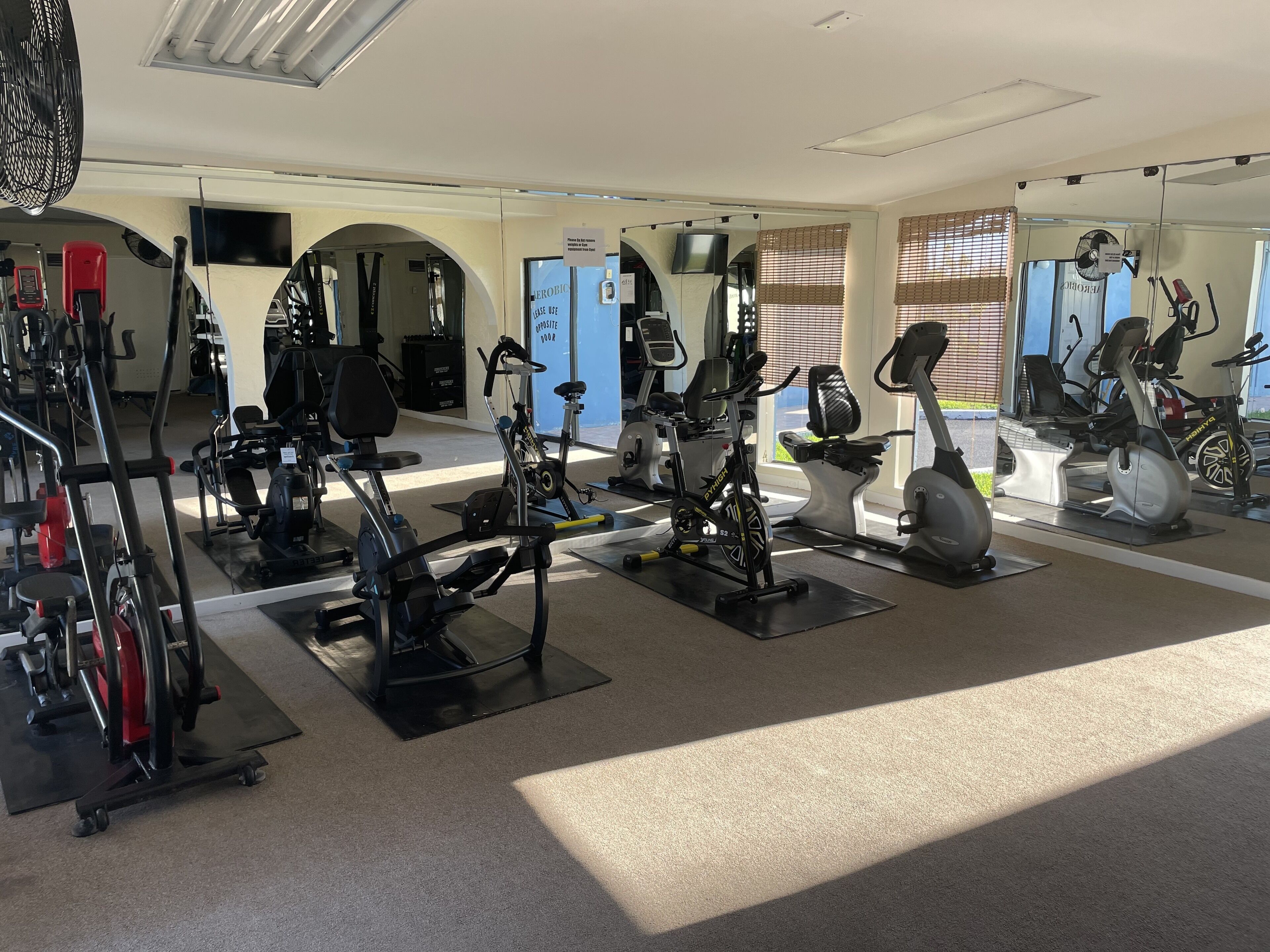 A gym with various exercise equipment, including stationary bikes and treadmills, arranged in a spacious layout with large windows allowing natural light.