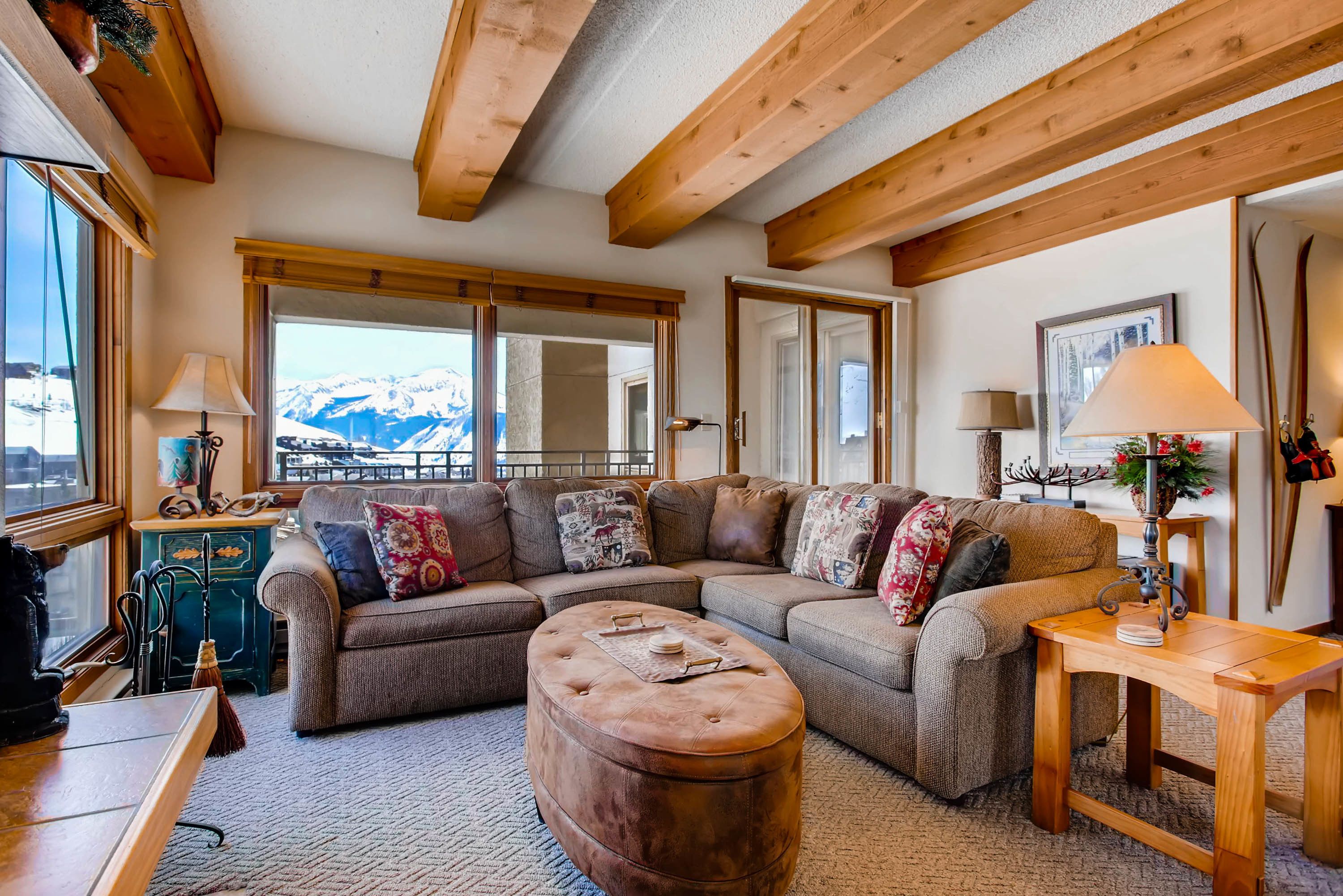 A cozy living room with a sectional sofa, ottoman, wooden beams, large windows with a mountain view, and a small table with a lamp.