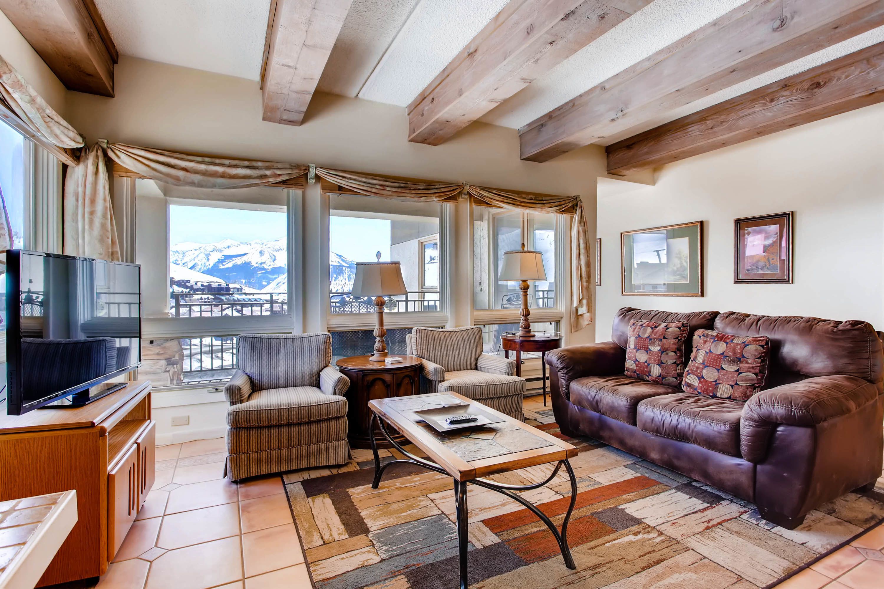 A cozy living room with a leather sofa, a television, and a view of snow-capped mountains through large windows.
