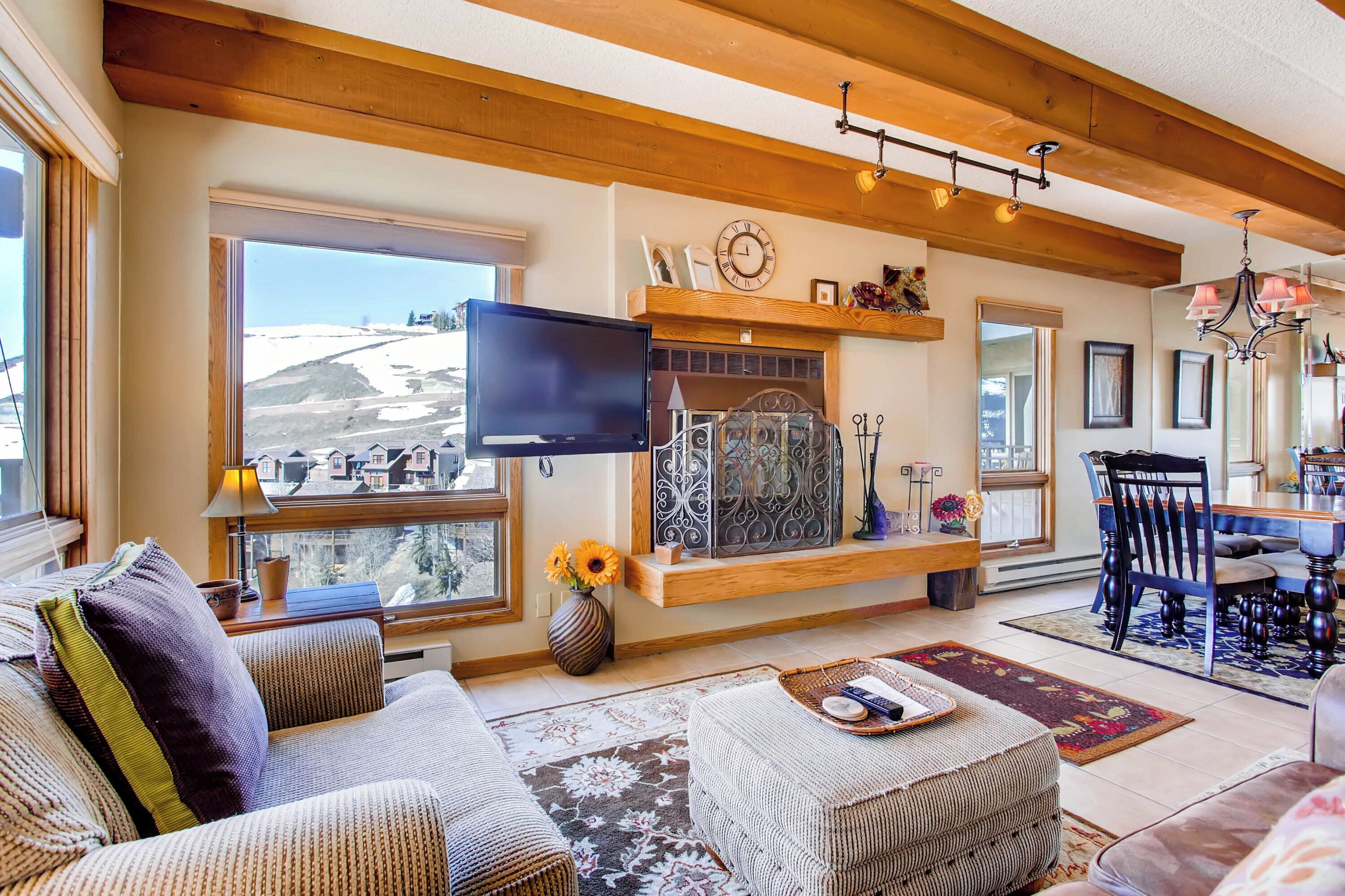 A cozy living room with a fireplace, a flat-screen TV, and a large window overlooking a snowy landscape.