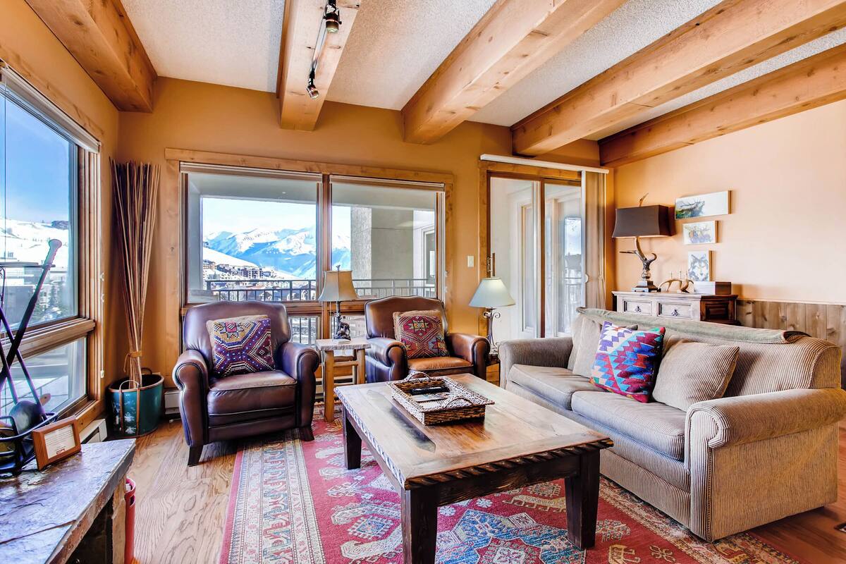 Un salon chaleureux avec un plafond en bois, de grandes fenêtres offrant une vue sur les montagnes, des canapés en cuir ornés de coussins à motifs et une table basse en bois.