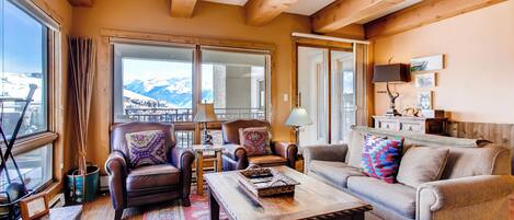 A cozy living room with a wooden ceiling, large windows with a mountain view, leather sofas with patterned cushions, and a wooden coffee table.