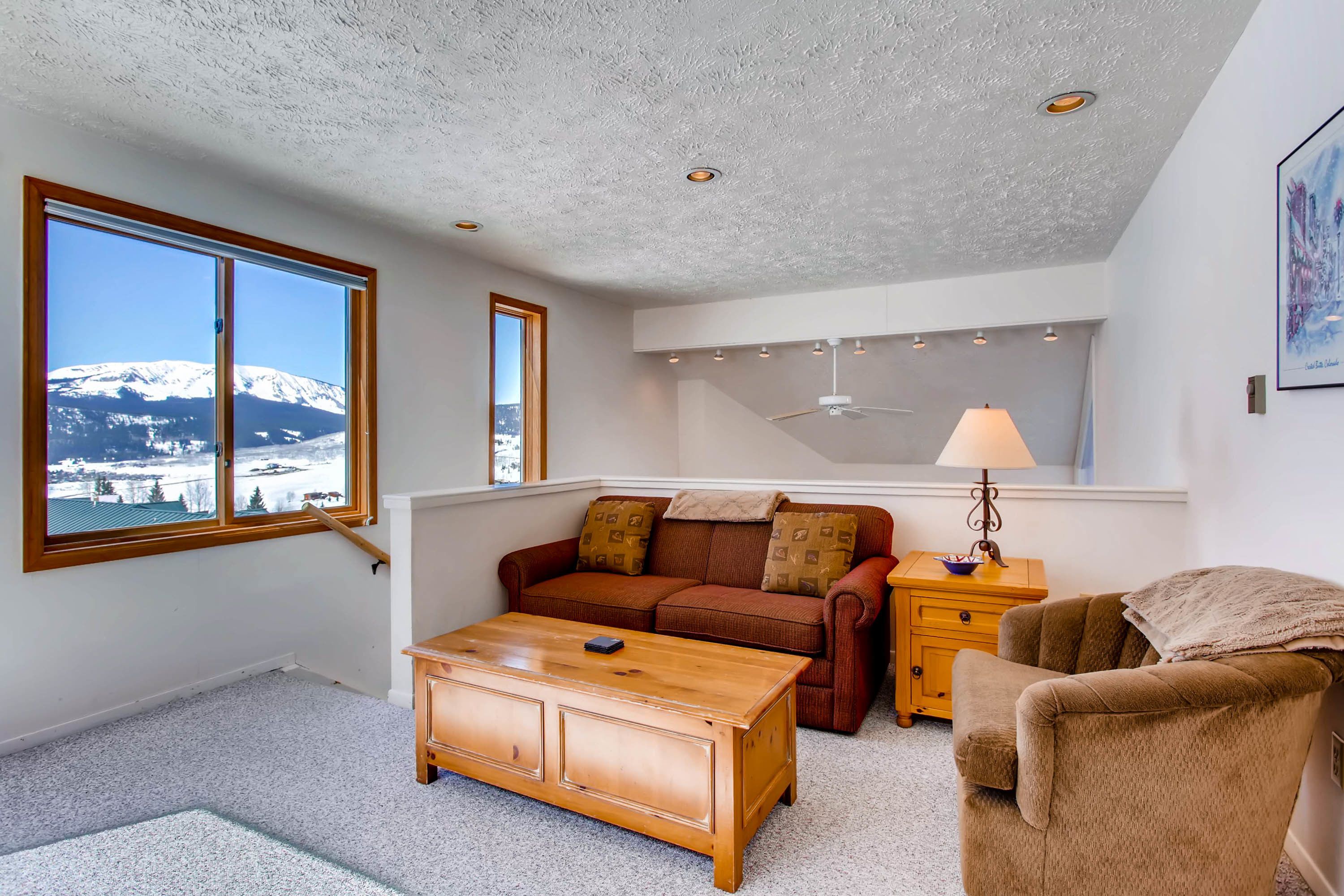 Una sala de estar acogedora con una mesa de centro de madera, un sofá marrón y un sillón color arena. Un amplio ventanal ofrece vistas a una montaña nevada.
