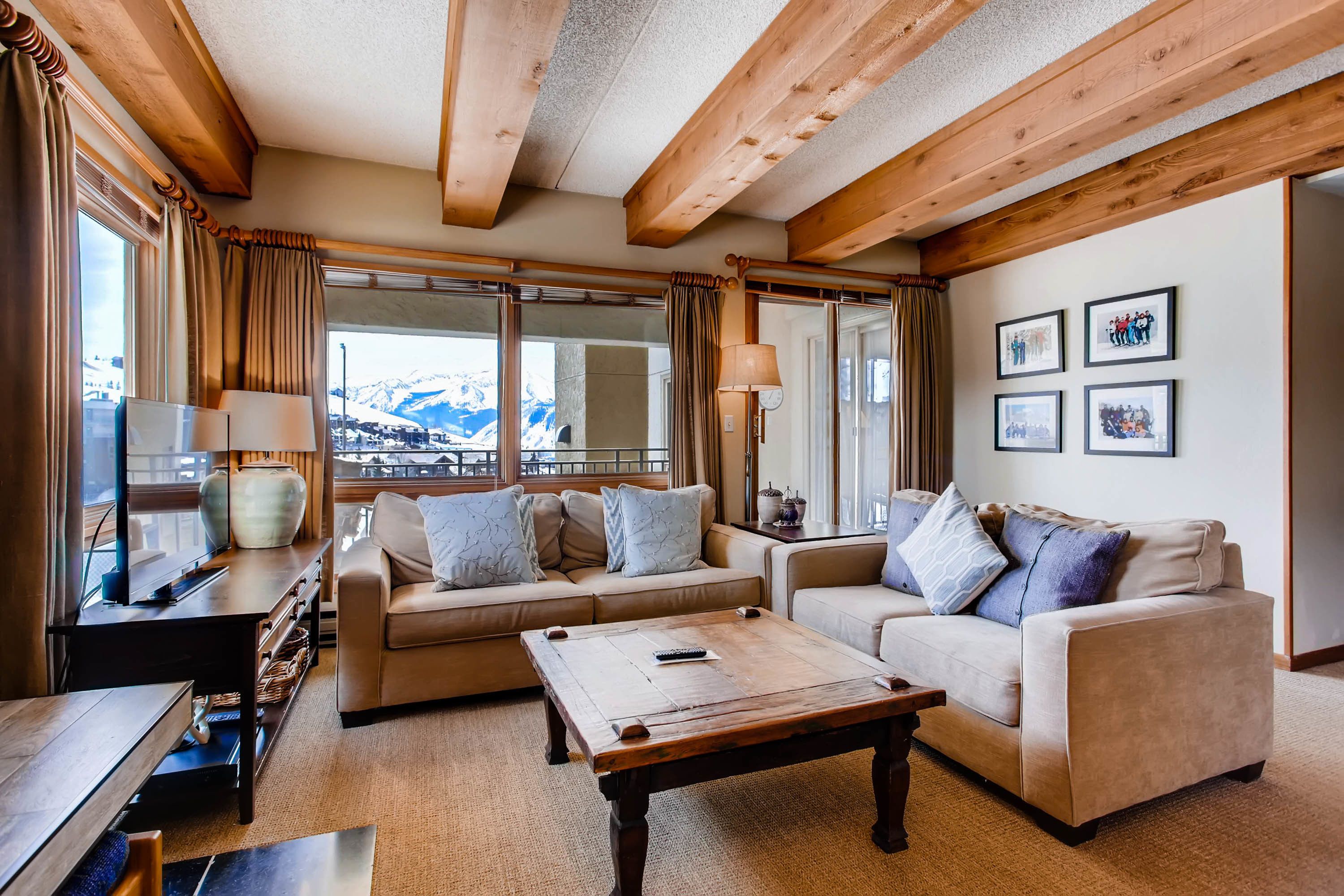 A cozy living room with two sofas, a coffee table, and a TV. Large windows offer a view of mountains.