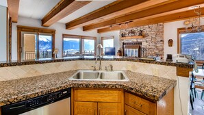 A kitchen with granite countertops, wooden cabinets, a stone fireplace, and large windows offering mountain views.