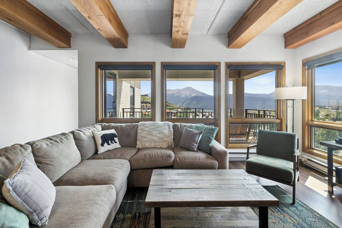 Un salon moderne avec un canapé d’angle, une table basse en bois et de grandes fenêtres donnant sur un paysage montagneux.
