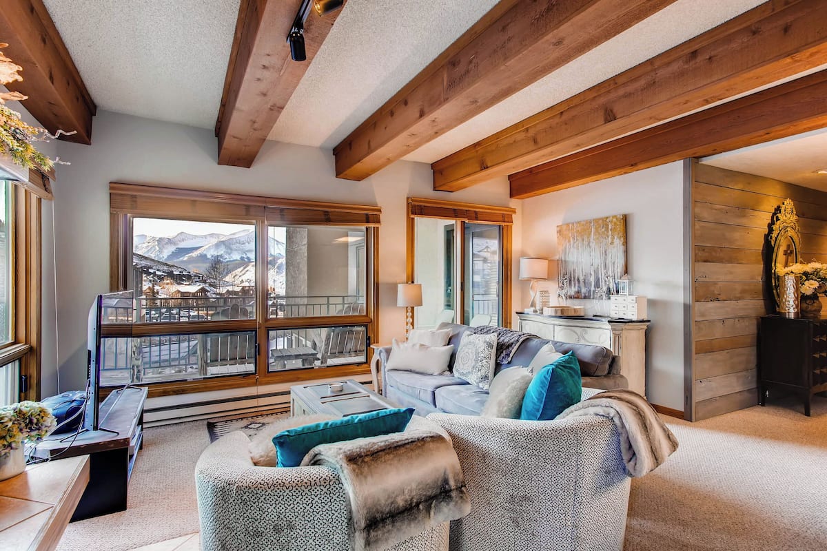 Un salon spacieux doté d’une grande fenêtre offrant une vue sur les montagnes, un coin salon confortable avec un canapé et des fauteuils, et un plafond en bois.