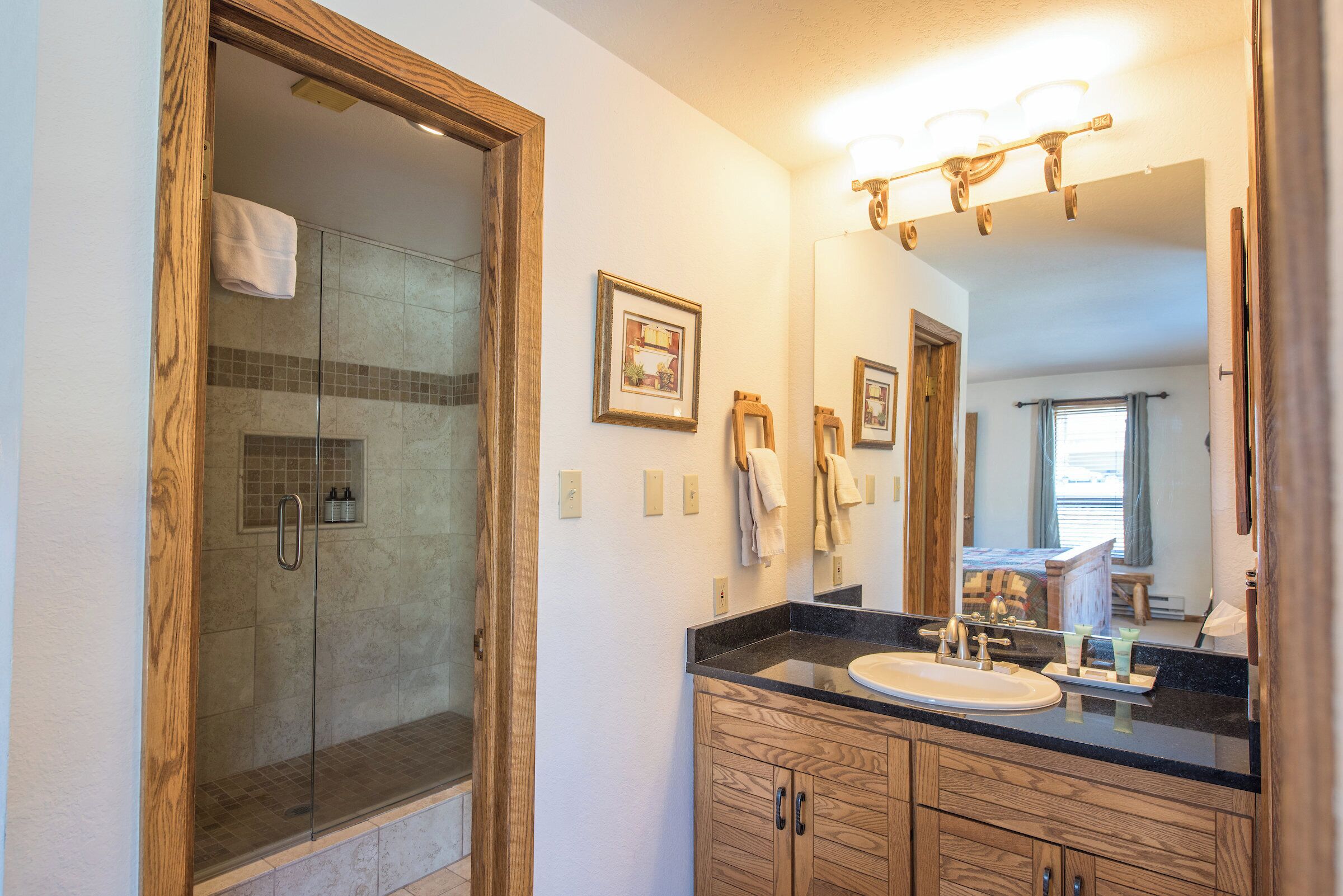 A bathroom with a glass-enclosed shower, a sink, wooden cabinets, and a mirror with lights above.