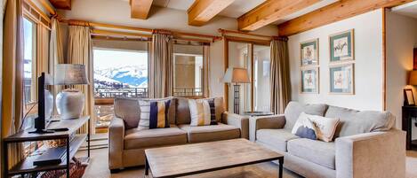 A cozy living room with two sofas, a coffee table, and a TV stand. Large windows offer a view of mountains.